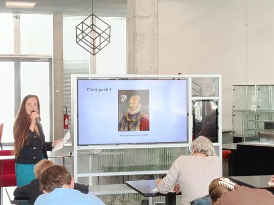 Laélia Véron parle au micro devant une salle, on voit des gens de dos qui écrivent des choses sur leurs pupitres. Au tableau blanc on voit une image de Montaigne
