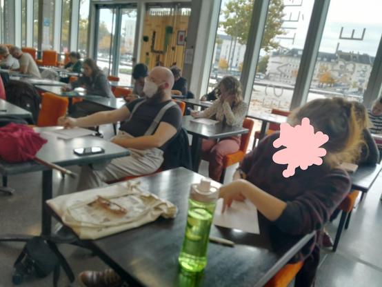 La même salle, on voit les gens de face, chaque personne à son pupitre. On ne distingue pas très bien les gens sur la photo, un visage au premier plan est caché