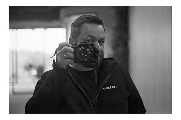 A black and white self-portrait of a man with a beard, wearing a dark zip-up hoodie, holding a Minolta film camera to his face.