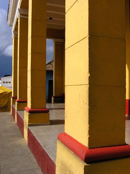 Five vertical brick columns shot so that the distance between them looks close. They are painted a dark yellow and red, and the setting sun is casting a golden glow on them which creates a strong contrast between light and shadow. In the distance are dark, stormy clouds. Trinidad, Cuba