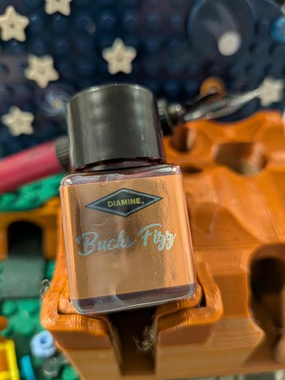 Dark blue Lego panel, with stars in background, in the foreground the bottle of Buck'sFizz ink dominates on the stand, while the dip pen leans in the midground.