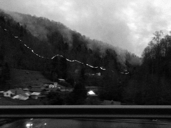 A grainy black and white photo taken at dusk showing a dark and heavily forested hillside as it slopes down to a small valley. A line of fire snakes through the frame and the middle of the hillside as clouds of smoke billow up in to the sky. A few houses with small patches of light are visible in the valley below the hill. A guardrail is prominent in the foreground.