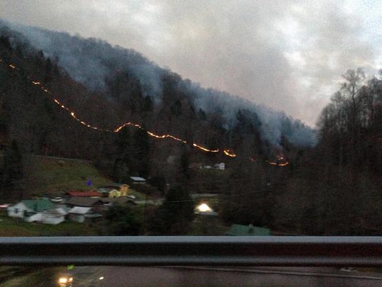 A grainy low-light photo taken at dusk showing a dark and heavily forested hillside as it slopes down to a small valley. A line of fire snakes through the frame and the middle of the hillside as clouds of smoke billow up in to the sky. A few houses with small patches of light are visible in the valley below the hill. A guardrail is prominent in the foreground.