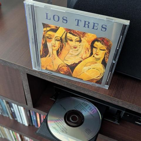 Una imagen muestra un álbum de música y un CD en un mueble de madera.

En la parte superior, un estuche de CD del álbum "Los Tres" está de pie. La portada presenta una pintura estilizada de tres mujeres con expresiones pensativas y vestimenta en tonos cálidos como amarillo, naranja y marrón. La mujer de la izquierda sostiene un abanico blanco, la del centro lleva un velo y la de la derecha apoya su mano en el pecho. El título "LOS TRES" se lee claramente en letras grandes, y "COLUMBIA" en el lomo.

Debajo, la bandeja abierta de un reproductor de CD revela el disco plateado del mismo álbum. En el CD se pueden leer el título "LOS TRES", "COLUMBIA" y algunos nombres de canciones como "Somos Tontos No Pesados" y "He Barrido el Sol". El entorno es un mueble de madera oscura con estantes llenos de otros discos a la izquierda y parte de un altavoz negro a la derecha.