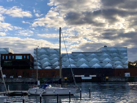 Vom Ufer der Schwentine auf das Geomar-Gebäude gesehen, im Schatten weiße Dreiecke auf der Fassade.  Davor auf dem Wasser ein Segelboot. Sonne steht niedrig so ist viel Schatten im Bild und viel blau.