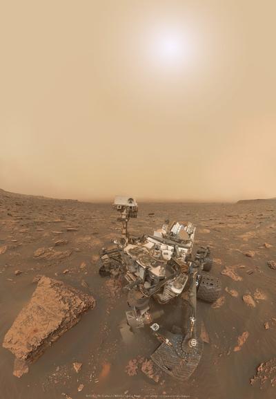 Sous un ciel aux teintes ocres uniformes, le Soleil perce au travers d'un halo diffus et un peu bleuté. Dessous, le rover Curiosity, véhicule autonome dont voit 4 des 6 roues, deux étant masquées par l'arrière du véhicule. En son coin gauche, un assemblage de caméras montées sur un mat. Beaucoup de câbles parcourent le corps du rover.
Le sol est composé de roches, avec de larges espaces remplis de sable. L'horizon est terne et peu discernable.