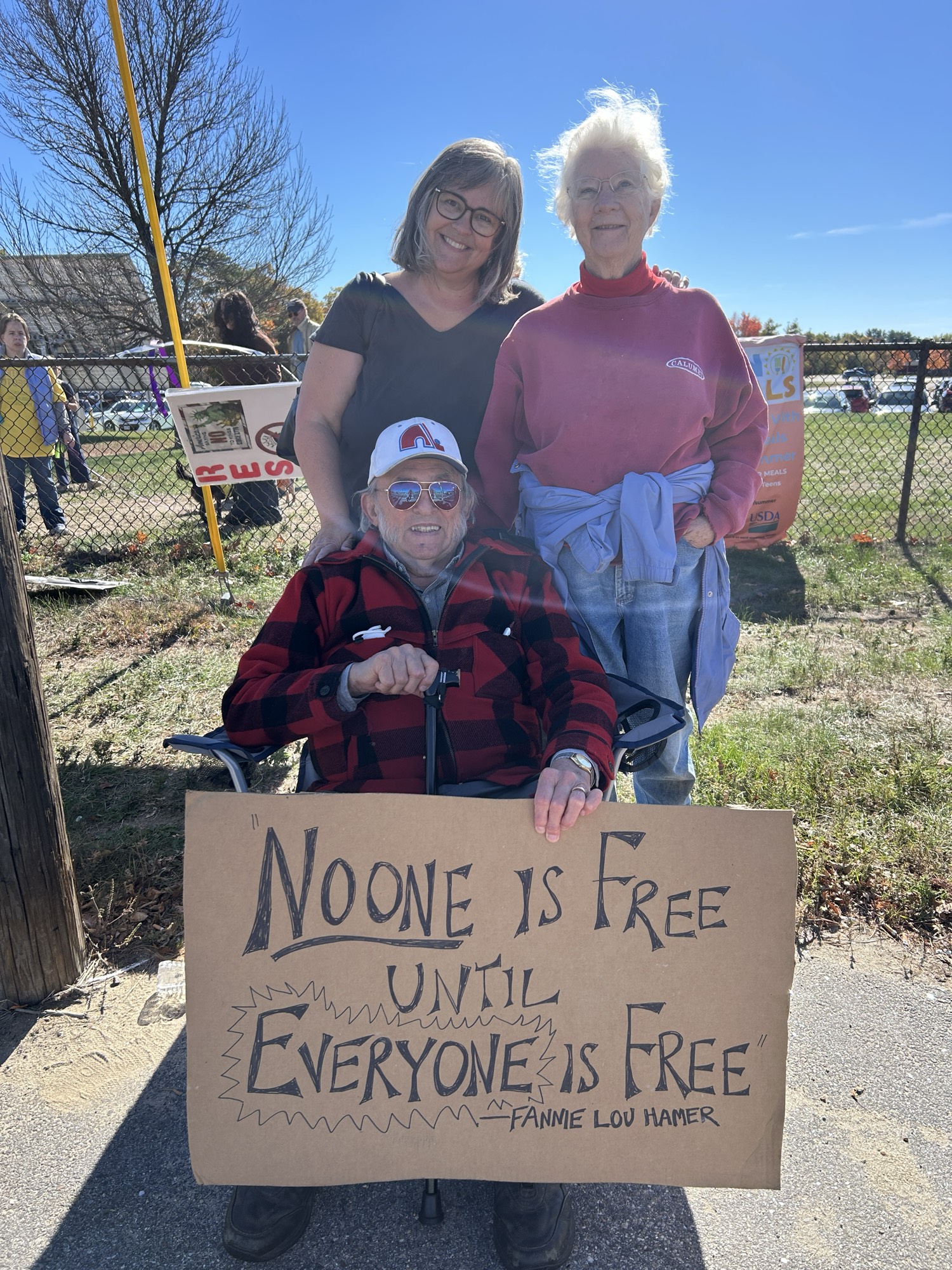 Three white adults, a middle-aged woman on the left, a woman in her 80s on the right, and a man in his 90s sitting in a chair wearing a baseball cap. In front of them is a cardboard sign that says “no one is free unless everyone is free — Fannie Lou Hamer.” It’s a bright sunny day with blue sky and they are smiling.