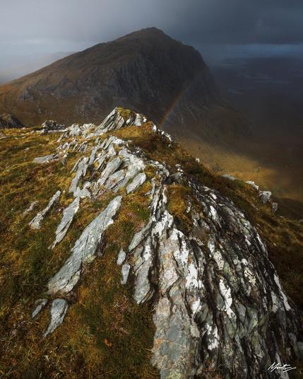 sharp sweeping gneiss rock comes in from the bottom right of the foreground leading down a mountain ridge. it is bathed in warm sunlight. the view leads out to a mountain summit in the dark shadow of an oncoming storm with a subtle rainbow coming in from the right hand side
