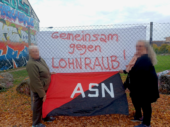 Zwei Gewerkschafterinnen stehen mit einer rot-schwarzen Fahne des ASN vor einem Banner: "Gemeinsam gegen Lohnraub!", das an einem Zaun hängt, im Hintergrund eine Hausfassade mit Graffiti
