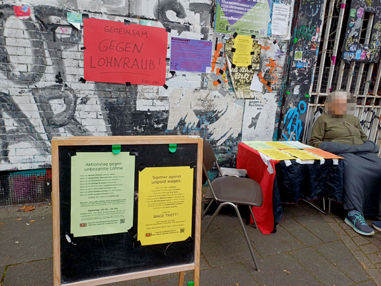 Ein Plakat-Aufsteller steht neben einem Infotisch an dem eine Person sitzt; im Hintergrund ein rotes Pappschild "Gemeinsam gegen Lohnraub!", das an einer HAusfassade mit Graffiti hängt