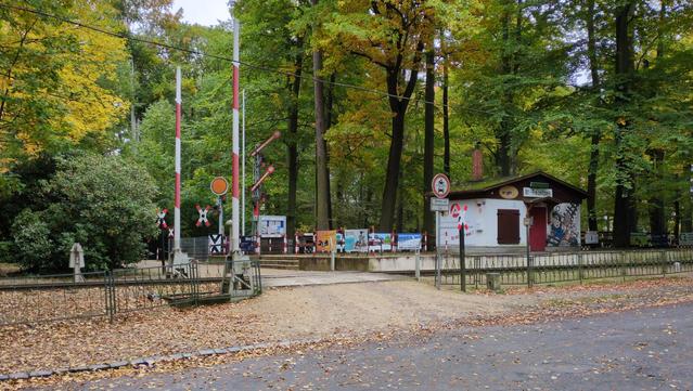 Stacja kolei wąskotorowej — parkowej. W centralnym punkcie przejazd kolejowy, przecięty jednym torem. Przejazd po obu stronach zamykają biało-czerwone rogatki, obie zamocowane po lewej stronie. Po prawej stronie znak zakazu wjazdu pojazdów silnikowych (motor i samochód w czerwonej obwódce) z tabliczką "Betriebs- und Versongungsdienst frei", a obok znak przejazdu kolejowego (dwie białe listewki z czerwonymi końcówkami, skrzyżowane pod wąskim kątem na dole i górze, a szerszym po bokach). Po bokach całość otacza zielony, metalowy płot. Za przejazdem, po prawej stronie peron z trzema stopniami schodów, a pośrodku budynek opisany "Bf – Freizeitpark", z czerwonymi drzwiami i oknem zamkniętym dwuskrzydłową, brązową okiennicą. Na prawo od drzwi mural przedstawiający uśmiechniętego, biegnącego po torach zawiadowcę z wielkim nosem, w niebieskim kostiumie, oraz rozkładającą powitalnie dłonie wiewiórkę. Peron otaczają biało-czerwone słupki z łańcuchem pomiędzy, w wielu miejscach wiszą plakaty z reklamami. Dalej kolejne znaki przejazdów, wskaźnik W1 i różne rodzaje semaforów kształtowych — najpewniej wystawowe. Całość otacza las liściasty, mieniący się zielenią, żółcią i pomarańczem.
