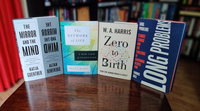 A photo of four books standing on a small, brown, wooden table. The out-of-focus background shows black shelves full of books, part of the beech-coloured laminate floor, and a red curtain covering the door.

From left to right, we have:
- The Mirror and the Mind, which divides its cover into a white and silver-coloured half and plays tricks by mirroring the title and author name;
- The Network of Life, showing a close-up of a leaf's vein pattern, the colours grading from yellow through green to blue;
- Zero to Birth, which shows a diagram of a human embryo inside the capital B of Birth, and the outline of a human brain in coloured dots that are connected by many lines;
- and Long Problems, which shows a dark blue background with the title in bold white letters, printed at a ninety-degree angle and narrowing towards the top, imitating the Star Wars title crawl.
