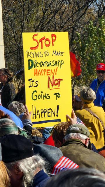 A close-up, slightly angled, outdoor shot depicts a protest sign held aloft by a group of people. The sign is a rectangular piece of yellow paper with black lettering. It reads: “STOP TRYING TO MAKE DICTATORSHIP HAPPEN! It’s NOT Going TO HAPPEN!”. The lower portion of the sign is partially obscured by the shoulders and backs of several people wearing colorful jackets and scarves. A red, white, and blue patterned item is visible in the foreground, and a person wearing a red cap is partially visible in the upper-right corner. The background appears to be a blurry landscape of trees and foliage.