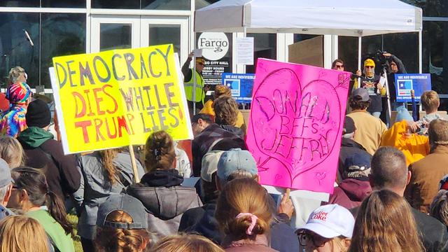 A crowd of people are gathered outside a building, likely a town hall, with signs protesting. Many individuals are holding brightly colored signs, with some partially visible in the foreground. A large yellow sign reads "DEMOCRACY DIES WHILE TRUMP LIES". Another sign, pink in color, displays the words “DONALD JEFFRY” in a handwritten style. The background features a building with glass doors and windows, a white tent, and other people standing.