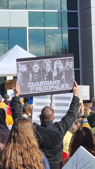A crowd of people are gathered outdoors, likely at a protest or demonstration, with a large sign being held aloft in the center of the frame. The sign is rectangular and features four grayscale portraits arranged horizontally. The text on the sign reads "GUARDIANS OF PEDOPHILES" in bold, capital letters. The person holding the sign is seen from the back, wearing a black jacket, and is surrounded by others in the crowd. The background includes a modern building with glass windows and a white tent.
