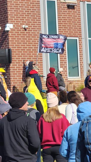 A crowd of people are gathered outdoors in front of a brick building with large windows. Several individuals are wearing hats and jackets, suggesting cool weather. In the foreground, people are close together, with backs to the camera, obscuring their faces and details of their clothing beyond color. A person in a red hat is holding a rectangular flag. The flag reads “NO KINGS IN AMERICA” in white capital letters against a striped background of red and white. The building's facade is made of red bricks and features dark green window frames.