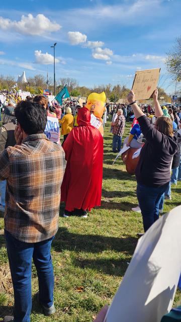 A photograph shows a crowd of people gathered on a grassy field under a clear blue sky. In the foreground, a person wearing a plaid shirt and jeans faces away from the camera, looking towards the center of the crowd. To the right of that person, another person is inflated in a red, Donald Trump-shaped costume. Further to the right, another person holds a white sign aloft, partially visible, along with other individuals holding various signs and papers. The background shows more people and a distant white building. 

The sign held aloft appears to have the text "No Parent Bridge". People are wearing a variety of clothing, including jeans, jackets, and plaid shirts. The grass is green and well-maintained, and the sky is a bright blue with a few scattered clouds.