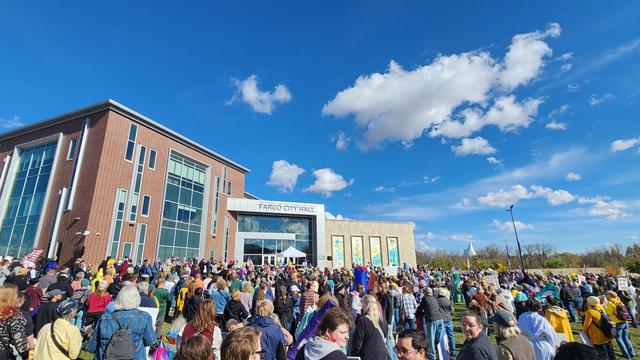 A large crowd of people is gathered in front of a modern brick building on a sunny day. The building has a glass facade and multiple windows, with a sign above the entrance reading “FARGO CITY HALL” in white capital letters. People are densely packed together, filling the foreground and extending back towards the building, with a variety of clothing colors visible. The sky is blue with scattered white clouds. In the lower left corner, a person is visible holding a sign, but the text is unreadable. The ground appears to be a mix of grass and paved areas, and trees are visible in the background.

Provided by @altbot, generated privately and locally using Gemma3:27b