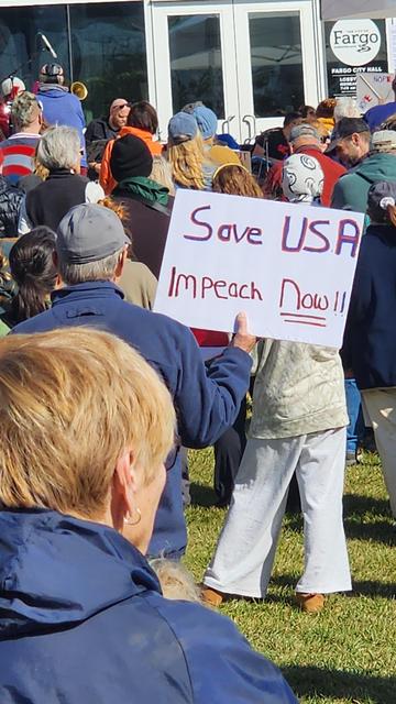 A crowd of people is gathered outdoors, likely at a protest or demonstration, as indicated by the sign being held aloft. The view is from behind a person wearing a blue jacket, focusing on the backs of heads and shoulders in the foreground. A person in a tan jacket is prominently holding a sign that reads "Save USA Impeach Now!!" in red and black lettering. The background shows a building with a sign that says "Fargo City Hall" visible on it. The scene appears to be well-lit, suggesting daytime, and there are numerous other people partially visible in the crowd.

Provided by @altbot, generated privately and locally using Gemma3:27b