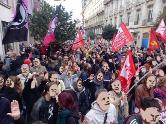 Vue en plongée d'une grosse foule dans la rue Jeanne d'Arc, arborant des drapeaux communistes et antifascistes.
Certain visage sont redessinés pour anonymiser