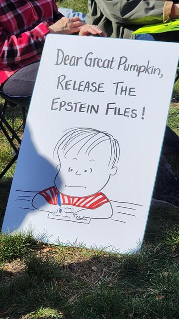A vertically oriented, close-up photograph shows a white, rectangular sign held outdoors on a grassy field. The sign has a simple, black-line drawing of a character’s face and torso, resembling a cartoon-style portrait. Below the drawing is text written in black marker. The text reads, "Dear Pumpkin, RELEASE THE EPSTEIN FILES!". The sign is slightly angled, and portions of people’s legs and clothing are visible in the blurred background. The sun casts a shadow on the sign.

Provided by @altbot, generated privately and locally using Gemma3:27b