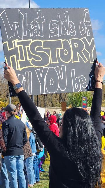 A crowd of people stands outdoors on a sunny day, holding signs and appearing to be at a protest or demonstration. The primary focus is on a large, rectangular sign held aloft by a person with dark curly hair. The sign is gray with large, white lettering that reads “What side of HISTORY will YOU be on?”. Several other people are partially visible in the background, wearing various clothing and holding additional signs. The scene suggests a gathering with a political or social message, with the central sign posing a question about participation in historical events.