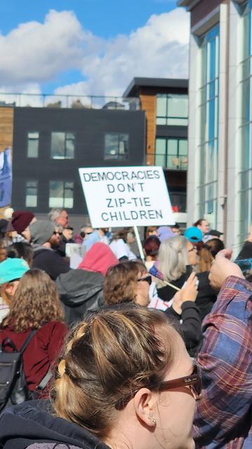 A close-up, outdoor photograph shows a large group of people gathered in what appears to be a protest or demonstration. The faces of several individuals in the foreground are visible, with varying hair colors and attire. A sign held aloft by a person in the crowd reads, "DEMOCRACIES DON'T ZIP-TIE CHILDREN." The background includes a building with large windows and a blue sky with some clouds. The overall scene suggests a politically charged event with a focus on children’s rights or welfare.