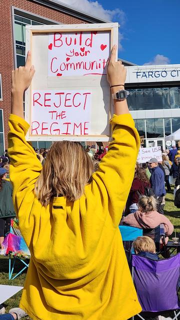 A person is holding a protest sign high in the air in front of a crowd of people and a building. The person is wearing a yellow sweatshirt and has their back to the camera. The sign is white with handwritten black text that reads “Build Your Community” and “Reject the Regime”. The crowd is partially visible behind the sign-holder and includes people of various ages sitting in folding chairs. The building in the background.