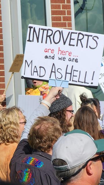 A close-up, waist-level shot depicts a crowd of people gathered, likely at a protest or demonstration. The focal point is a large, handmade sign held up in front of a storefront window. The sign is white with black lettering that reads, “INTROVERTS are here… and we are MAD AS HELL!” Several individuals are visible in the foreground, partially obscuring the view of others further back in the crowd. One person in the foreground is wearing a denim jacket with patches, and another has on a gray baseball cap. The window behind the sign appears to be displaying various items, and a glimpse of a green sign can be seen on the right edge of the frame.