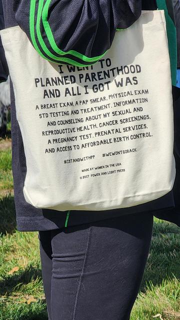 A white tote bag is shown hanging from someone's shoulder, partially obscuring a person wearing a dark-colored patterned shirt. The bag features text in black capital letters that reads: "I WENT TO PLANNED PARENTHOOD AND I GOT WAS A BREAST EXAM, A PAP SMEAR, PHYSICAL EXAM, STD TESTING AND TREATMENT, AND COUNSELING ABOUT MY SEXUAL AND REPRODUCTIVE HEALTH, CANCER SCREENINGS, A PREGNANCY TEST, PRENATAL SERVICES, AND ACCESS TO AFFORDABLE BIRTH CONTROL." Below the list of services are the hashtags "#ISTANDWITHPP" and "#WEWONTGOBACK". At the bottom of the bag, the text "MADE BY WOMEN IN THE USA" and "©2017 POWER AND LIGHT PRESS" are visible. The background is blurry, featuring green foliage.

Provided by @altbot, generated privately and locally using Gemma3:27b