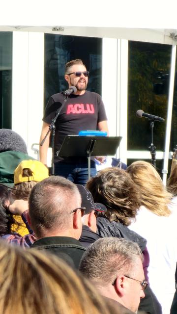 A person is speaking at a podium in front of a crowd, likely outdoors. The speaker is standing behind a dark-colored podium with a music stand holding papers. The speaker is wearing sunglasses and a black t-shirt with the text "ACLU" in red capital letters across the chest. The crowd is visible in the foreground, partially obscuring the view, with multiple heads and shoulders filling the frame. The background is a white structure, possibly a trailer or tent, with dark windows.

Provided by @altbot, generated privately and locally using Gemma3:27b