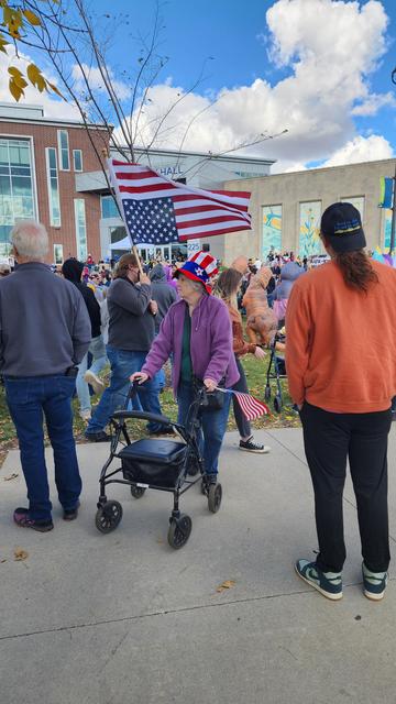 A crowd of people are gathered outside a brick building with a green roof, likely during an outdoor event. A person in a purple jacket is slowly walking forward while pushing a rolling walker, and appears to be the focus of the image. Several people are holding American flags in the background, and others are standing around watching. The building has a sign that reads "HALL" visible on its facade. The sky is blue with some clouds, and fallen leaves are scattered on the ground.

Provided by @altbot, generated privately and locally using Gemma3:27b