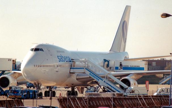 Front side view of a very large, 4 engined jet airliner parked facing to the left, with a white set of mobile airstairs with a large awning over the stairs connected to the forward mid-cabin door on the planes left side - the airstairs have blue "Sabena" titles on the side.
The plane is almost all white, with blue "Sabena" titles on the upper forward fuselage, between to 2 passenger decks.
The white tail has a dark blue circle containing a stylised, elongated letter "s".
A white truck has its rear raised up to the rear door, the truck body being white, with a blue stripe, a black stick figure holding a brush and a bucket, and the black words "Cabin Cleaning" giving the purpose of this vehicle away.
Various other airport vehicles are scattered around the plane, including a low-slung blue tug attached to the nose wheel.
Bright but hazy sky fills the rest of the frame.