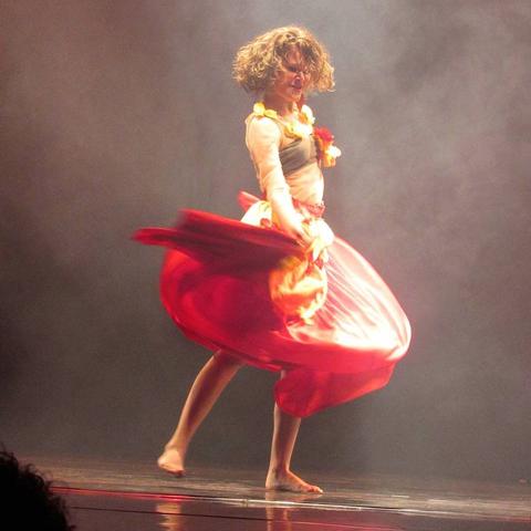 Uma dançarina, descalça sobre um palco, de cabelo tamanho médio, loiro, ondulado, vestindo uma roupa de tecido transparente e saia longa vermelha, segurando uma parte da barra e a agitando no ar.