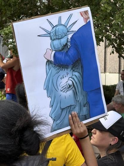 Protester at a “No Kings” rally in Orlando holds a sign showing Donald Trump hugging the Statue of Liberty, who is making the international hand signal for HELP behind her back, with the word “HELP” written in large letters next to her