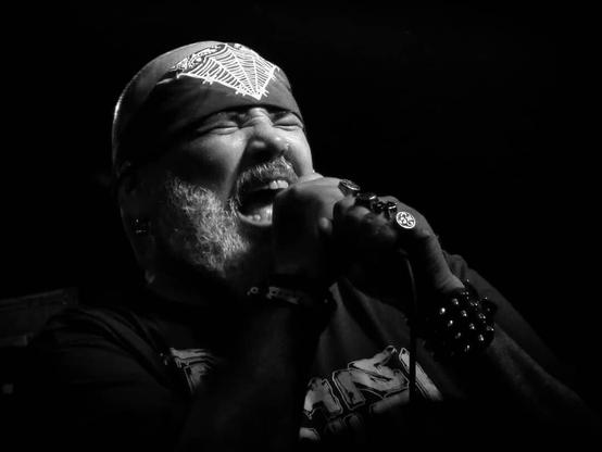 Fotografia em preto e branco do vocalista da banda Ratos de Porão, João Gordo, segurando o microfone com as duas mãos, de olhos fechados, durante um show e "gritando". Ele tem uma bandana sobre a testa, usa uma pulseira com espinhos e diversos anéis nos dedos.