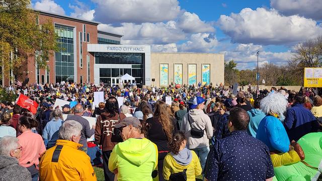 A large crowd of people is gathered in front of a building with the text “FARGO CITY HALL” visible above the entrance. The people are densely packed, facing toward the building, and many are holding signs. Individuals in the crowd are wearing a variety of clothing in different colors, including yellow, blue, and black. The building is a modern structure with a glass facade and vertical windows, and has two large murals on its side. A small sign to the right reads “PROJECT 2040”.

Provided by @altbot, generated privately and locally using Gemma3:27b