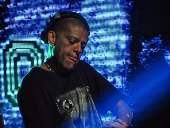 KL Jay discotecando durante o show. Ele é um homem negro com a pele com algumas manchas de vitiligo, tem a cabeça raspada, está com um headphone sobre a cabeça, mas fora da orelha e está com a língua parcialmente para fora (sinal de esforço). Ao fundo um grande painel digital com partes de uma imagem não identificável.