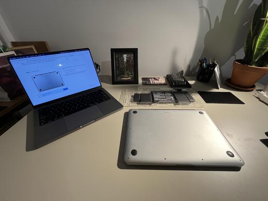 Two Macbooks on a desk, one showing repair instructions, another face down ready to be opened up. Behind the new battery and tools.