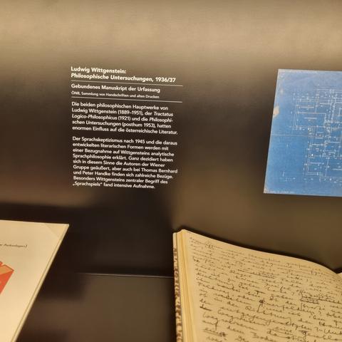 A museum plaque stating in German that this is Wittgenstein Philosophical Investigations manuscript. A corner of the manuscript can be seen.