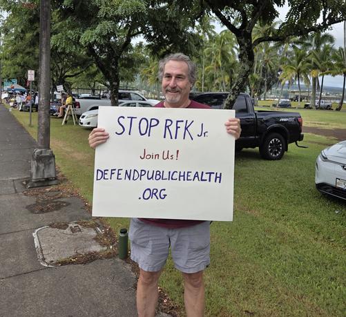 Older man holding a sign reading "Stop RFK Jr.  Join Us! defendpublichealth.org"