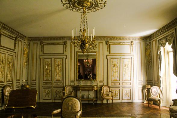 A wide shot of various chairs and neoclassical furniture in a sunlit room. The wall panels and doors are cream with gold borders.