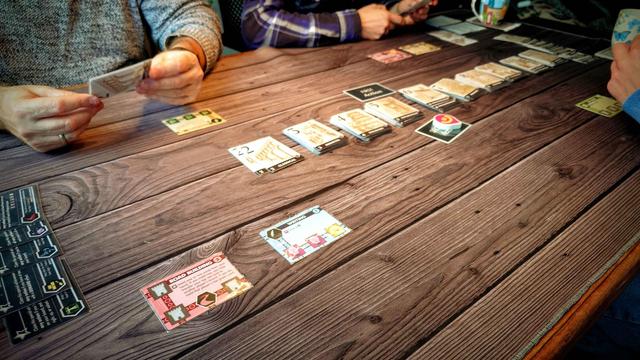 Ein Tisch mit einer frisch angefangenen Partie Innovation. Drei Spieler bzw ihre Hände und Arme, die Karten halten sind angeschnitten zu sehen. In der Mitte des Tisches verdeckte Kartenstapel