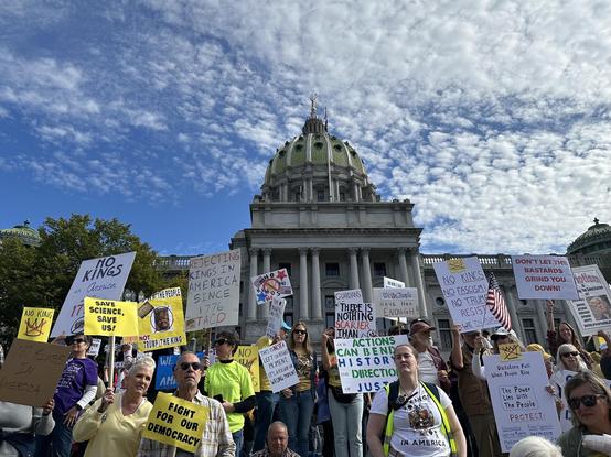 A small crowd of crowd of No Kings protesters begins to gather before the official start of the protest. They stand on the steps of the Pennsylvania State Capitol building, Harrisburg PA, facing the street and showing their No Kings signs — most of them hand printed. It’s about an hour before noon on a sunny October day. A glittering gold statue tops the green Capitol dome under the bright blue sky with mackerel clouds.