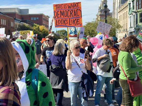 A lively crowd of No Kings protesters stand, walk, and talk in the blocked-off street that crosses in front of the Pennsylvania State Capitol building, where more protestors gather on and around the steps. A protester wearing a green terry cloth pajamas is on the left. In the center, a woman holds a ‘This is what Democracy Looks Like’ sign while glancing at her cell phone. A pink and white inflatable unicorn is behind her, in a fairly dense crowd that is waving signs, listening to speakers, and talking.