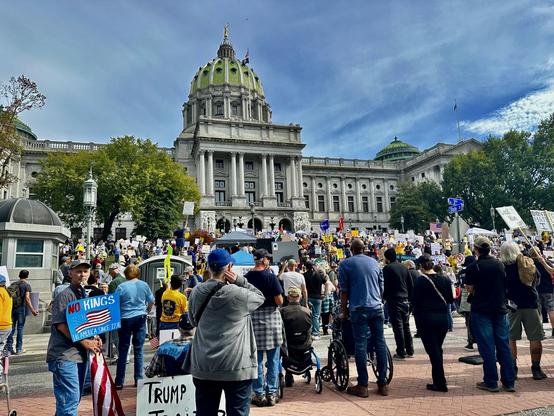 A line of No Kings protesters — including three in wheel chairs — line the street facing the Pennsylvania State Capitol building, Harrisburg PA. The protesters in the street and on the Capitol steps carry a wide variety of homemade and printed No Kings signs. It’s past noon on a sunny October day. A glittering gold statue tops the green Capitol dome, but the blue sky darkens as a thin layer of clouds approach.