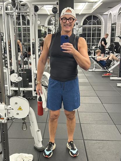 A man is taking a selfie in a gym. He is wearing a sleeveless black shirt, blue shorts, and a white cap. A water bottle and towel are in his hands. The gym is visible behind him, featuring exercise equipment and other gym