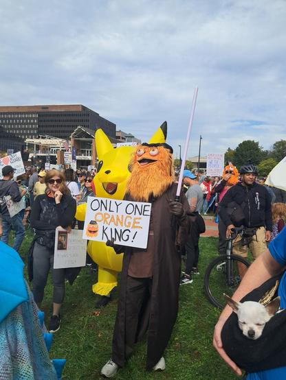 Protesters are gathered on the grass in Philadelphia, Pennsylvania, USA. One person is dressed in a Gritty costume. Gritty is furry and orange, and he the mascot of the Philadelphia Flyers. He is holding a sign that reads, "Only One Orange King."