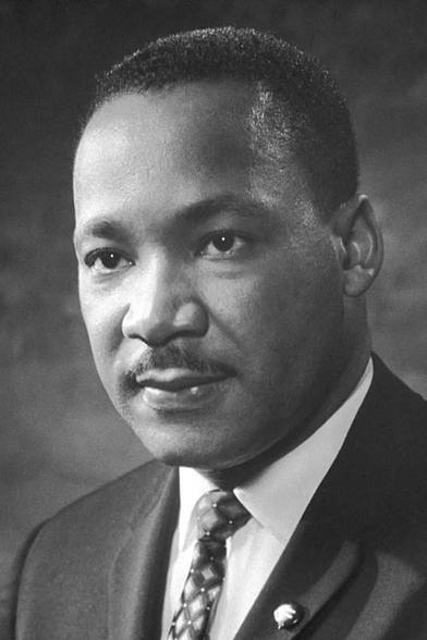 Martin Lither King Jr in 1964. He's a Black man with short copped black hair and a narrow moustache. He's wearing a dark suit and tie, and looking intently off camera with a serious expression.