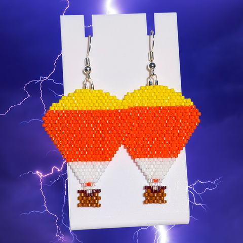 A pair of beaded candy corn hot air balloon earrings over a purple lightning background.